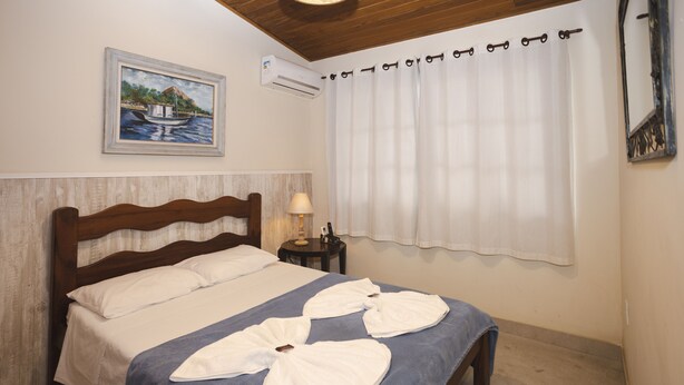 A neatly made bed with white linens, a wooden headboard, and a painting of a boat on the wall. - R. Virgínia 48 (Búzios)