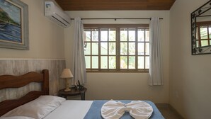 A hotel room with a bed, a window with curtains, a painting on the wall, and an air conditioning unit. - R. Virgínia 48 (Búzios)