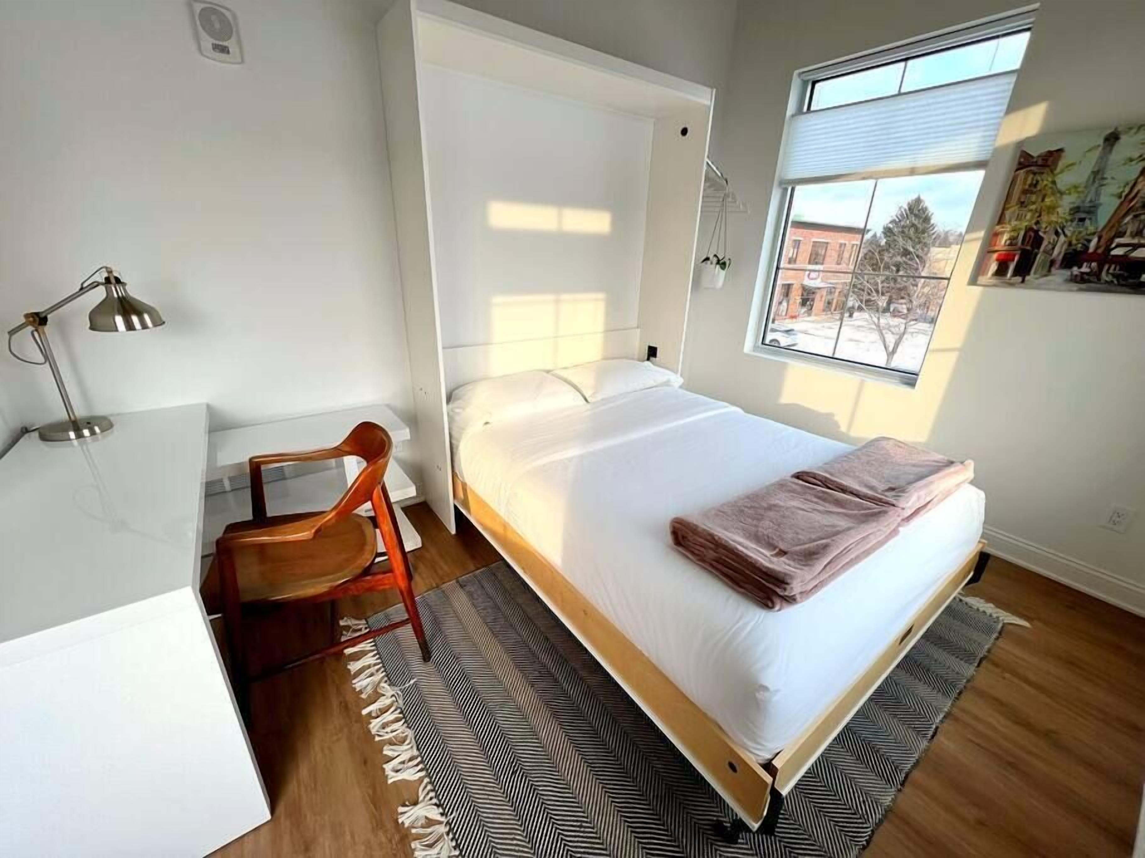 A bedroom with a bed, a desk, a chair, a lamp, and a window with a view of buildings.