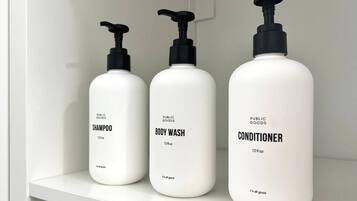 Three white bottles of public goods shampoo, body wash, and conditioner on a white shelf.