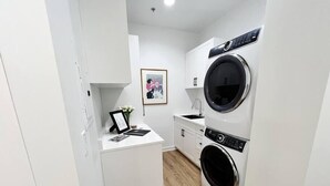 A compact laundry room with a front-loading washer and dryer, a small sink, and a framed picture on a white counter. - The Quarters | Suite 1 (Grafton)