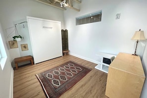 A room with a wooden floor, a patterned rug, a white door, a bench, a small table, a lamp, and a shelf with a plant.