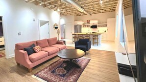 A spacious living area with a pink sofa, a round coffee table, and a patterned rug. A kitchen with modern appliances is visible in the background. - The Quarters | Suite 3 (Grafton)