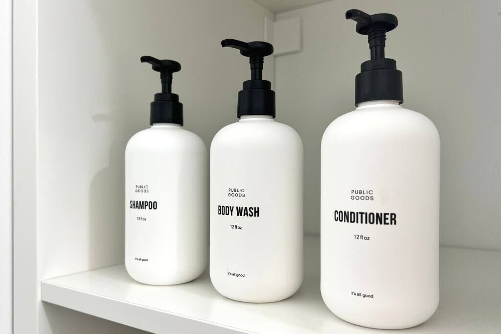 Three white bottles of public goods shampoo, body wash, and conditioner on a white shelf.