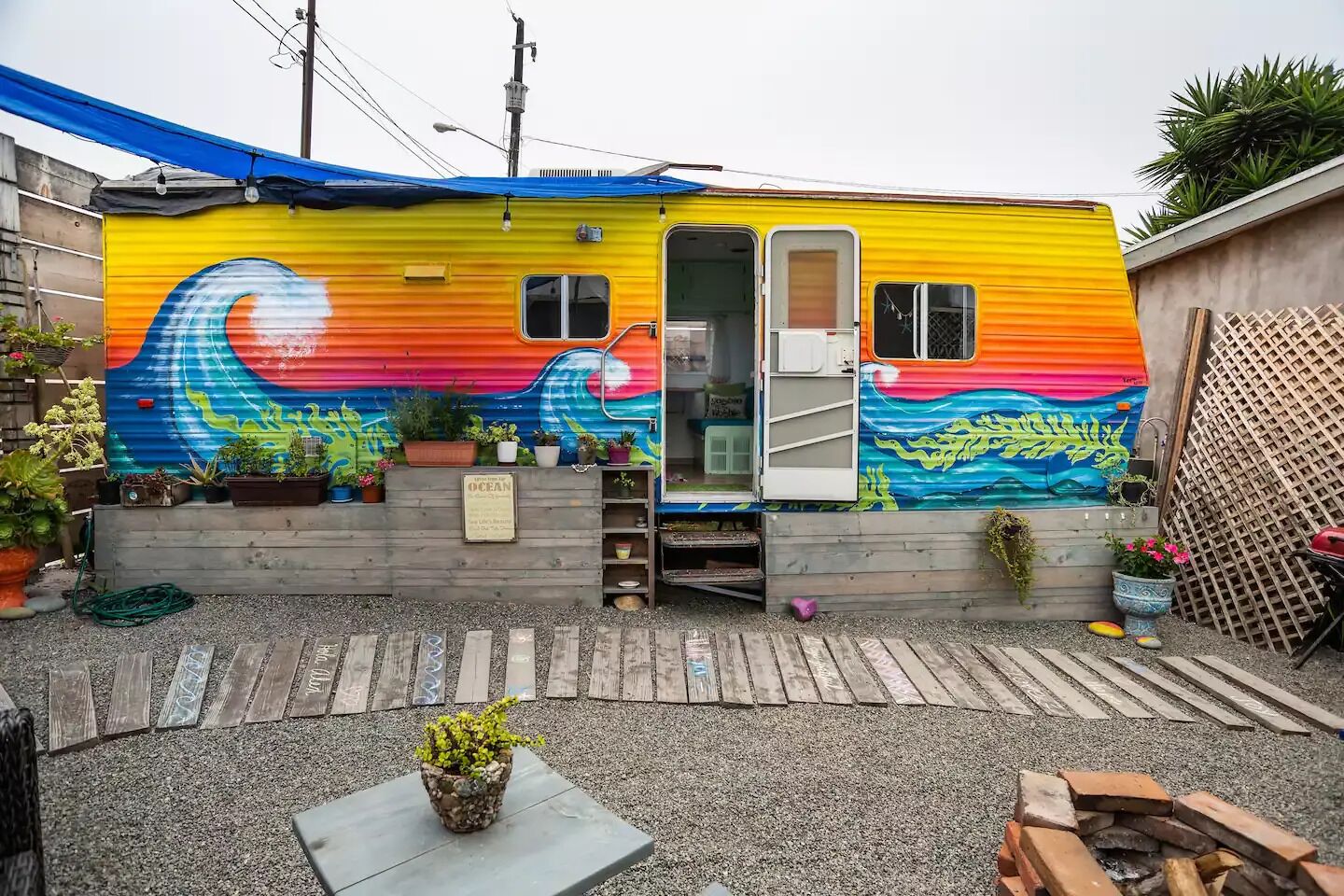 Uma casa móvel colorida com um mural vibrante de ondas, um deck de madeira e diversos vasos com plantas.
