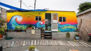 Ein farbenfroher Wohnwagen mit einem lebhaften Wellen-Mural, einer Holzdielen-Terrasse und verschiedenen Topfpflanzen.