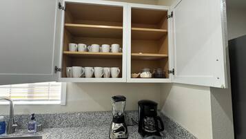 A kitchen with a granite countertop, a coffee maker, a blender, and a cabinet with white mugs and jars.