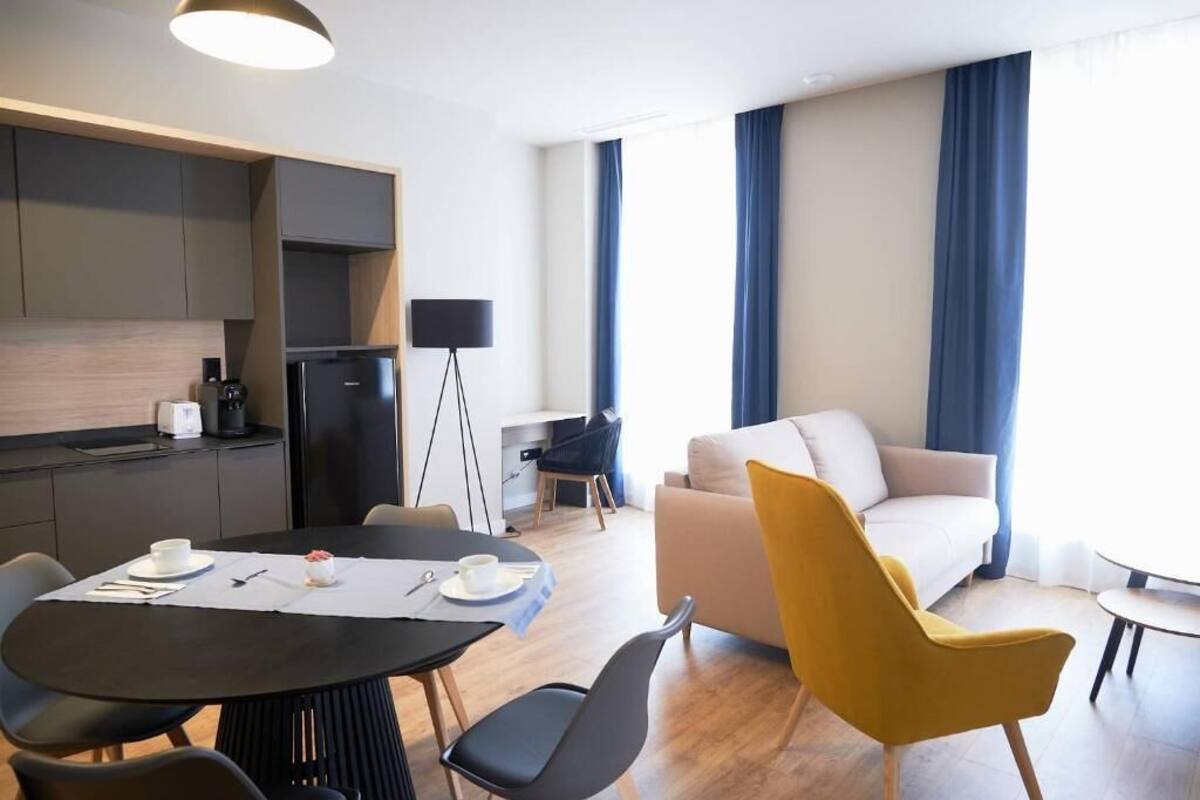 A modern hotel room with a kitchen area, a dining table with chairs, a sofa, and a coffee table.
