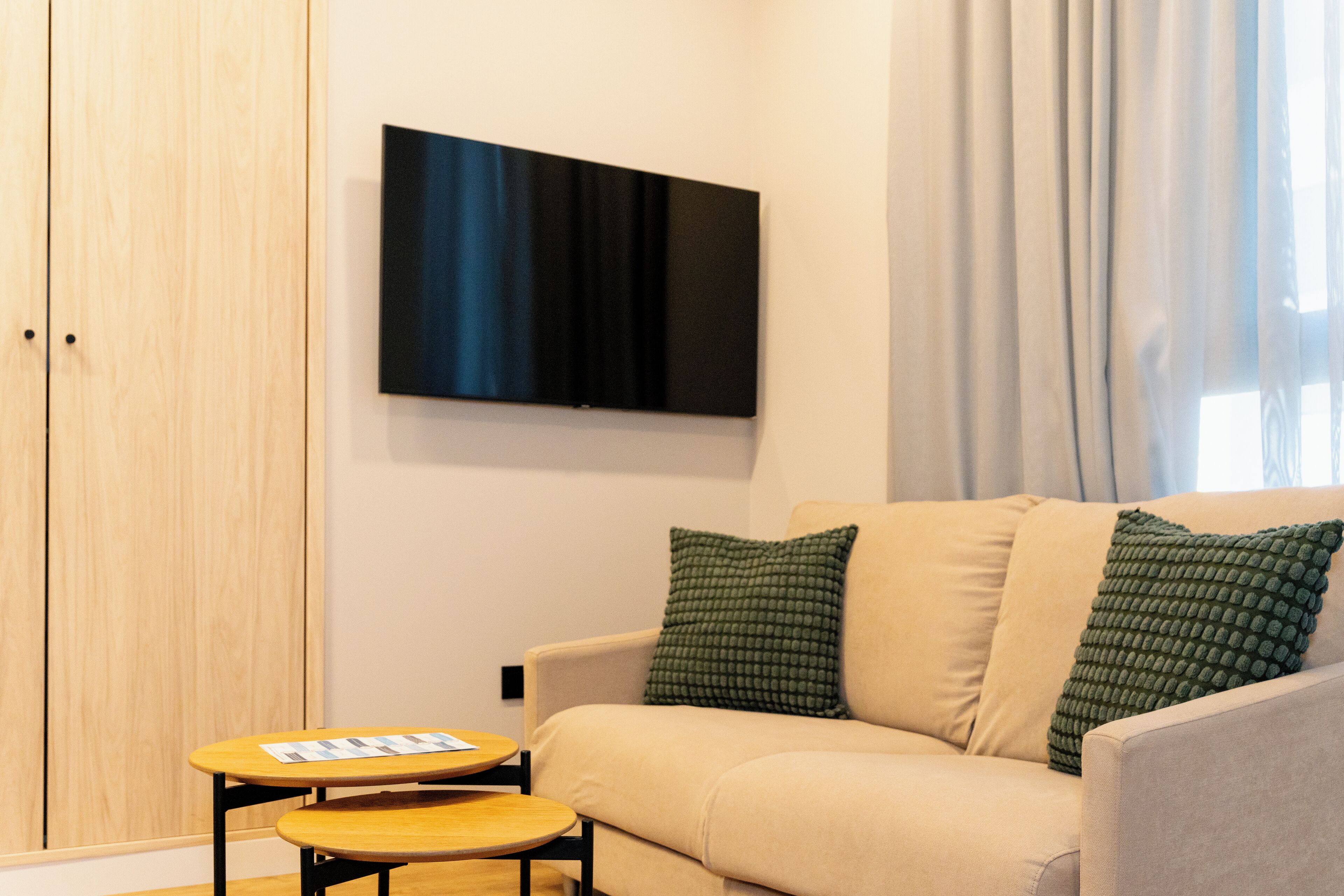 A modern living room with a beige sofa, two green pillows, a flat-screen TV on the wall, and a small wooden table with a magazine.