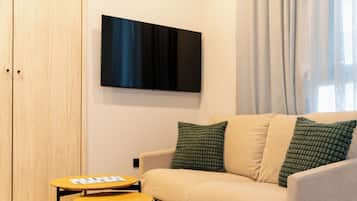 A modern living room with a beige sofa, two green pillows, a flat-screen TV on the wall, and a small wooden table with a magazine.