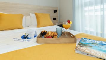 Habitación de hotel con una cama, una bandeja con cruasanes, un vaso de zumo de naranja y una revista sobre la cama.