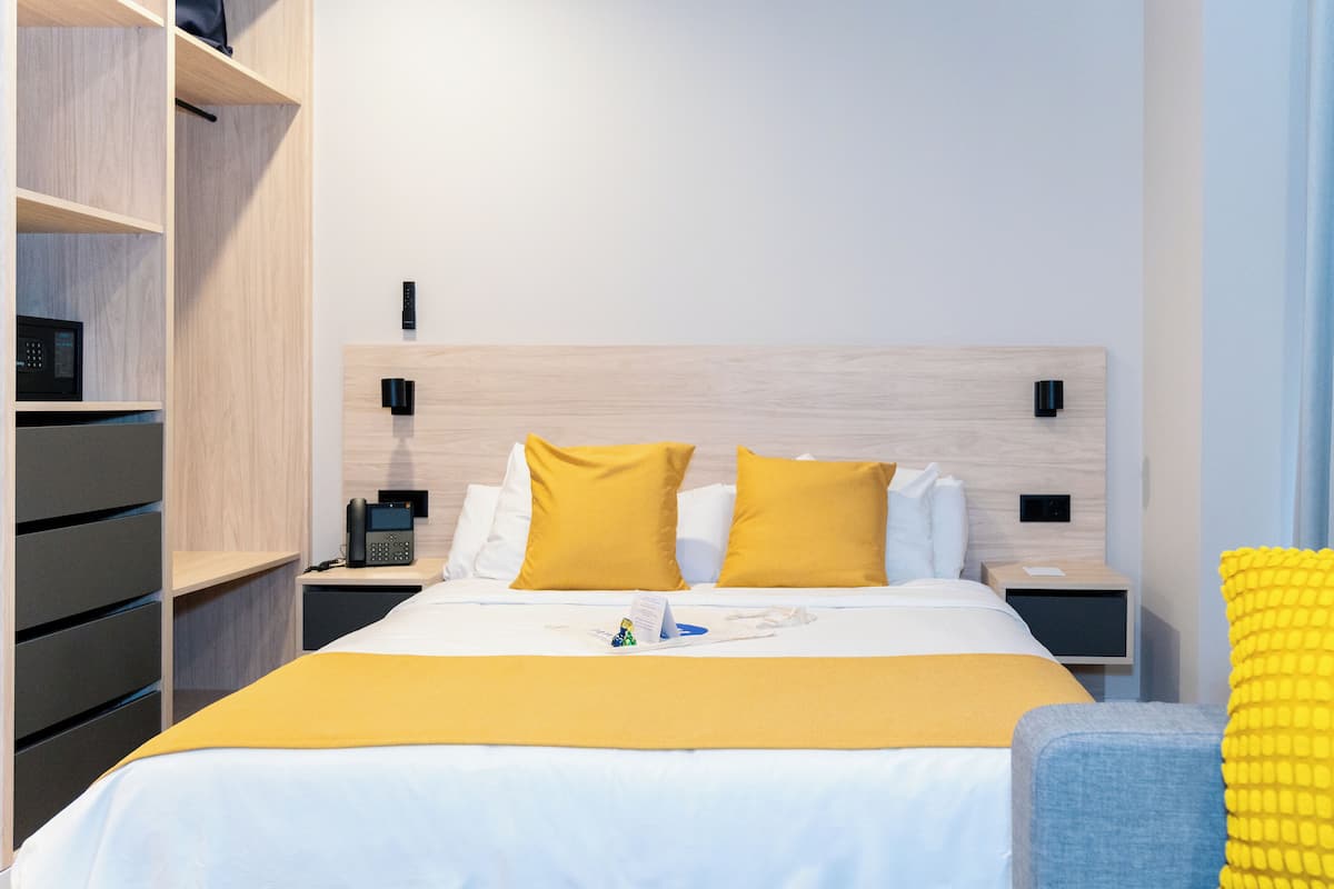 A modern hotel room with a wooden headboard, a bed with white and yellow bedding, a bedside table with a phone, and a nightstand with a lamp.