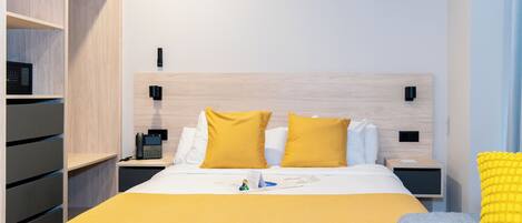 A modern hotel room with a wooden headboard, a bed with white and yellow bedding, a bedside table with a phone, and a nightstand with a lamp.