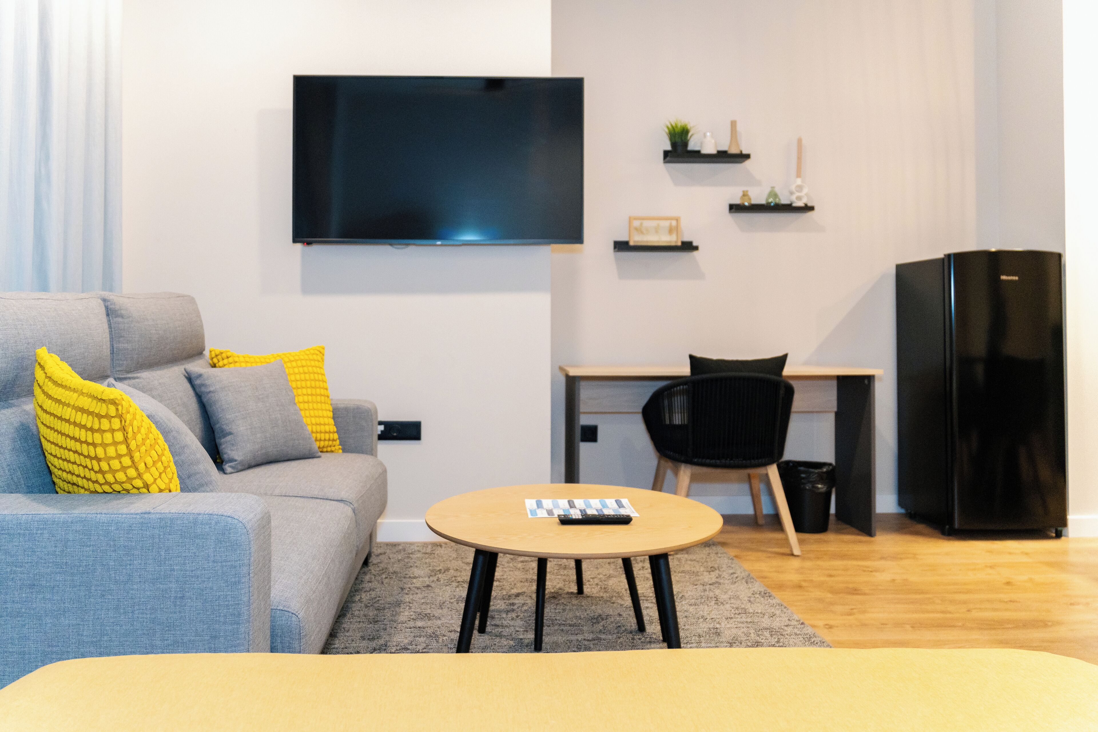 Una sala de estar moderna con un sofá gris, cojines amarillos, una mesa de centro redonda, un televisor de pantalla plana, un escritorio pequeño, una silla negra y un refrigerador negro.