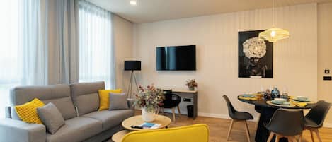 A modern living room with a grey sofa, yellow armchair, dining table set for four, and a flat-screen TV mounted on the wall.