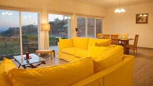Ein helles Wohnzimmer mit einem gelben Sofa, einem Esstisch mit Stühlen und großen Fenstern mit Blick auf einen Hügel.