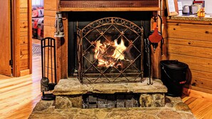 L’intérieur chaleureux d’un chalet avec une cheminée en pierre, des murs en bois, un téléviseur fixé au-dessus de la cheminée et un coin cuisine sur la droite.