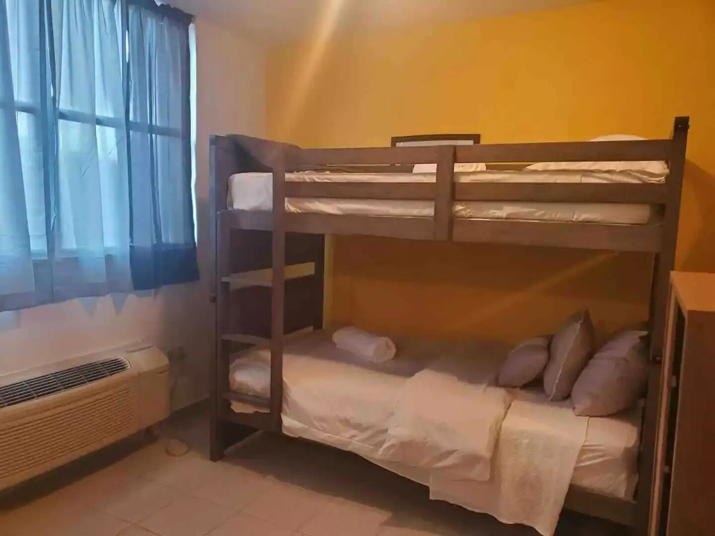 Een slaapkamer met een stapelbed, raam, airconditioning en een houten bureau.