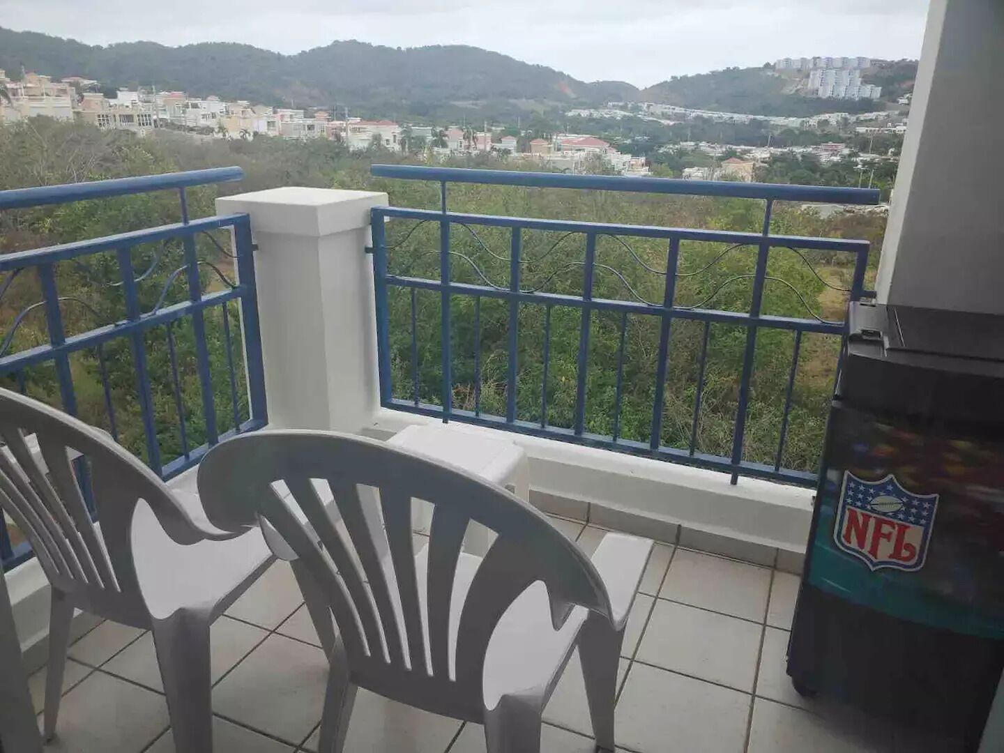 A balcony with a view of a cityscape, featuring a railing and two white plastic chairs.