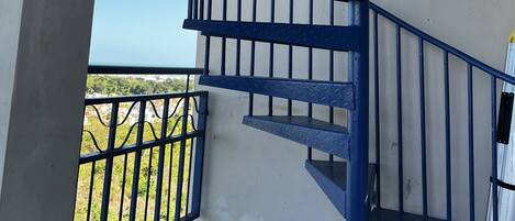 Una escalera espiral azul con barandilla de metal que conduce a un balcón exterior con vistas a zonas verdes y edificios.