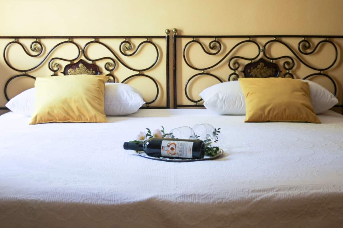 Een opgemaakt bed met een decoratief hoofdeinde, twee kussens en een fles wijn op het beddegoed.