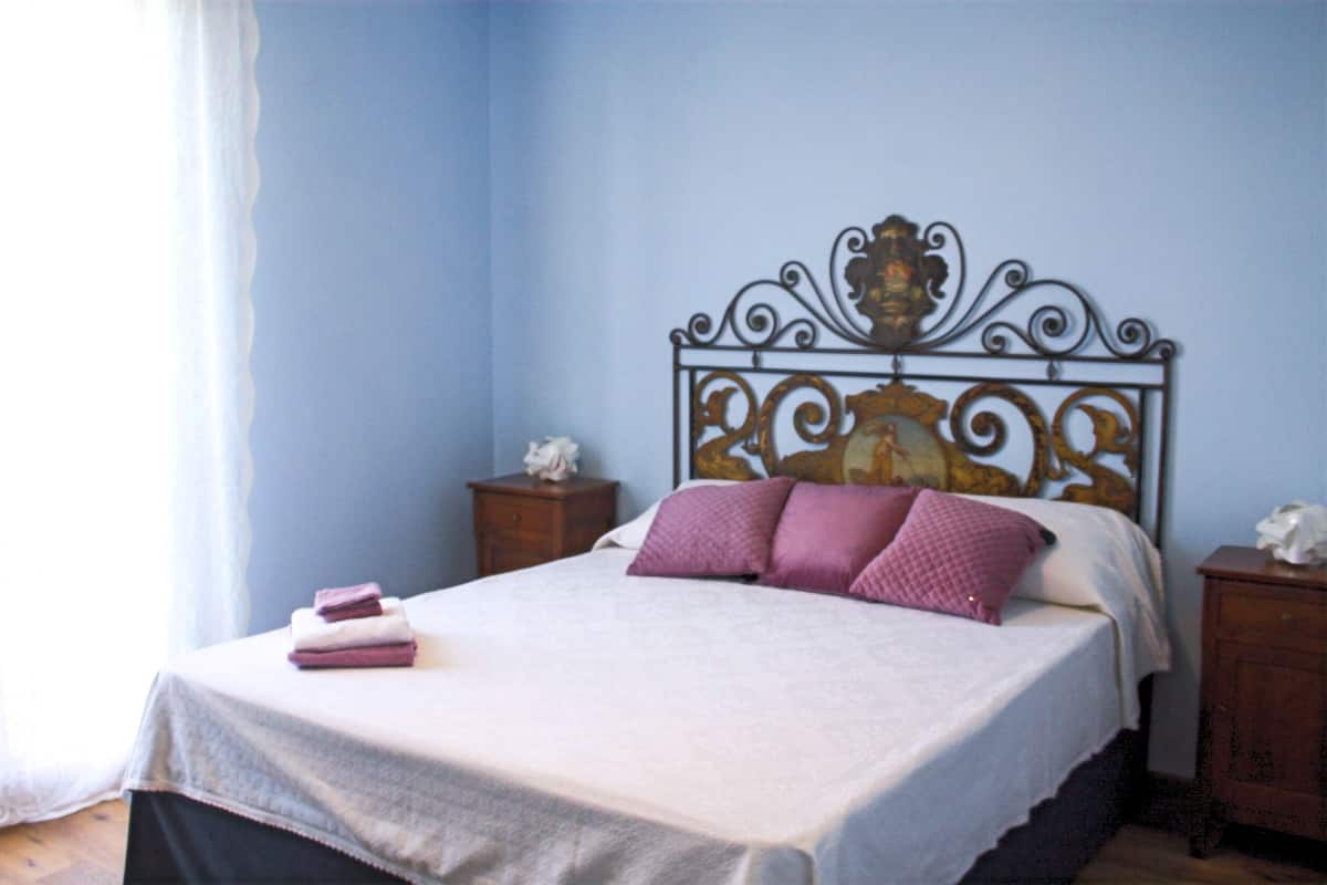 Een netjes opgemaakt bed met een witte dekbedovertrek en roze kussens, een houten nachtkastje met een zakdoekendoos, en een decoratief metalen hoofdbord.