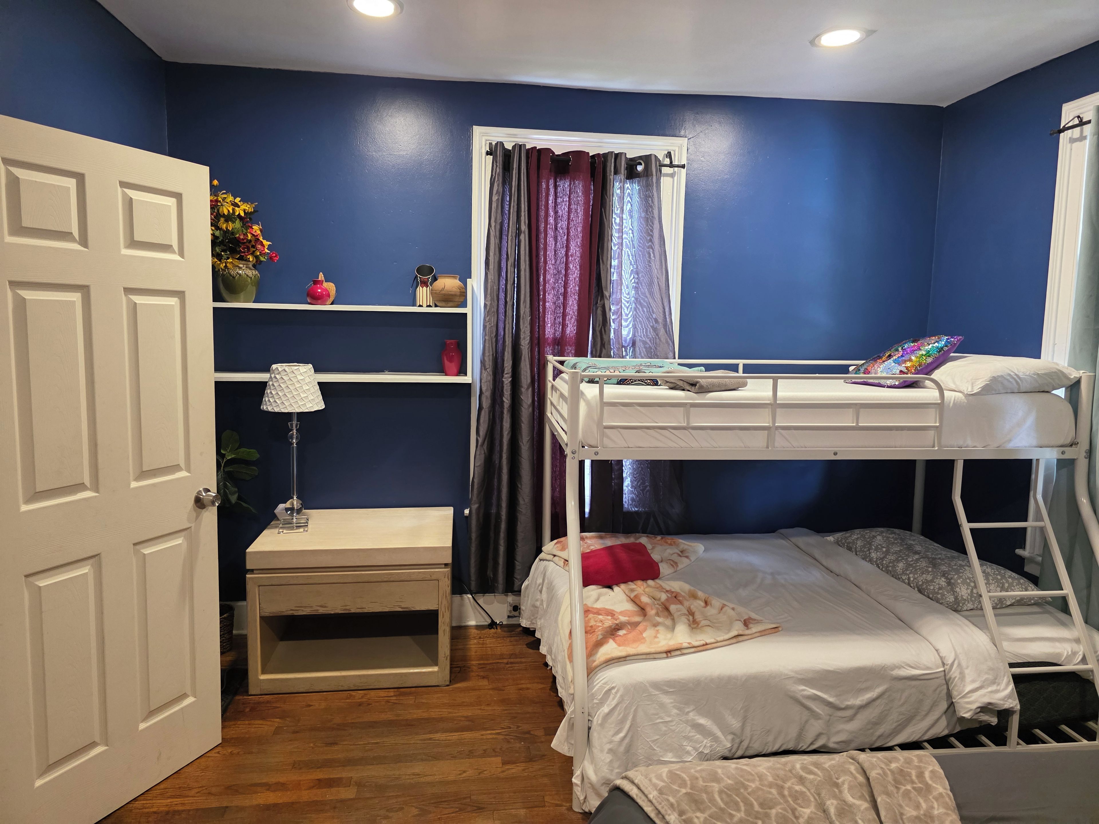 Une chambre à coucher avec un lit superposé, une table de chevet, une lampe, une étagère avec des objets décoratifs et une porte.