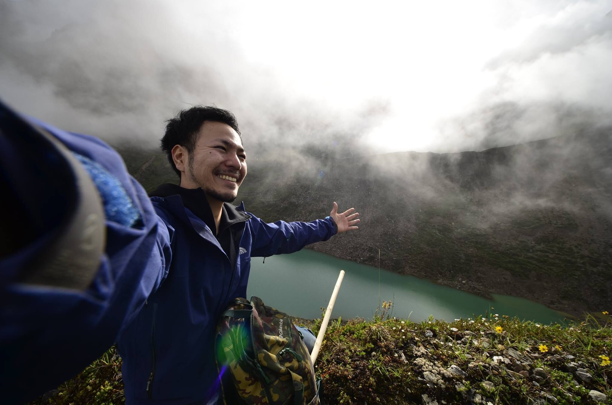 A person in a blue jacket standing with arms outstretched, overlooking a lake with a mountainous backdrop and mist.