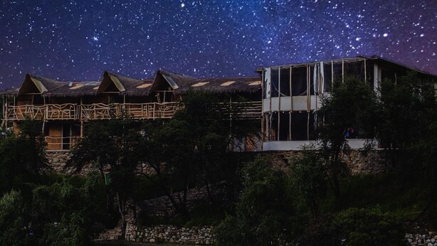 A starry night sky over a rustic building with thatched roofs and a modern structure with large windows. - Refugios Salkantay (Mollepata)