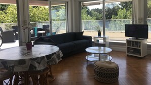 A spacious living area with a dark-colored sofa, a round dining table with a patterned tablecloth, a small white side table, a round ottoman, and a television on a white stand. Large windows offer a view of greenery outside. - Arcachon Pereire Appartement Pieds Dans Leau (Arcachon)