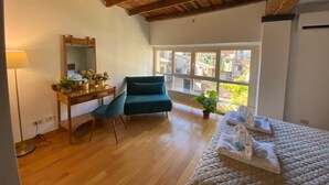 Una camera da letto con soffitto in legno, un letto, una scrivania, una sedia, una finestra vista edifici e una pianta in vaso.
