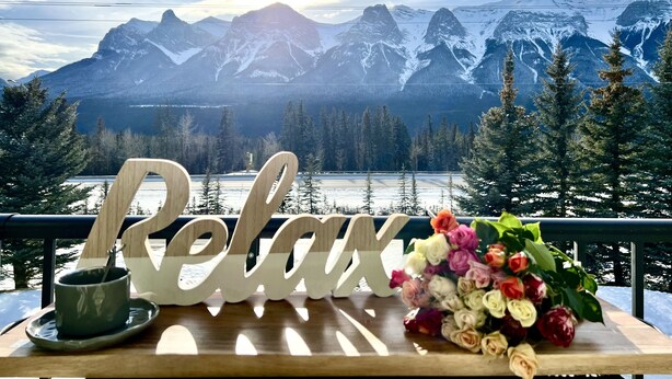 A wooden table with a 'Relax' sign, a cup, and a bouquet of flowers, overlooking a mountainous landscape with a lake and snow-covered peaks.