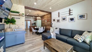 A modern living room with a wooden ceiling, a gray sofa, a dining area with a table and chairs, and a kitchen visible through an open door.
