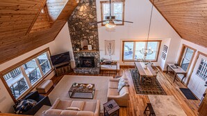 A spacious loft-style living area with a stone fireplace, wooden ceiling, and large windows offering views of the outdoors.