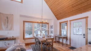 A spacious dining area with a wooden table and chairs, a chandelier, and large windows providing natural light.