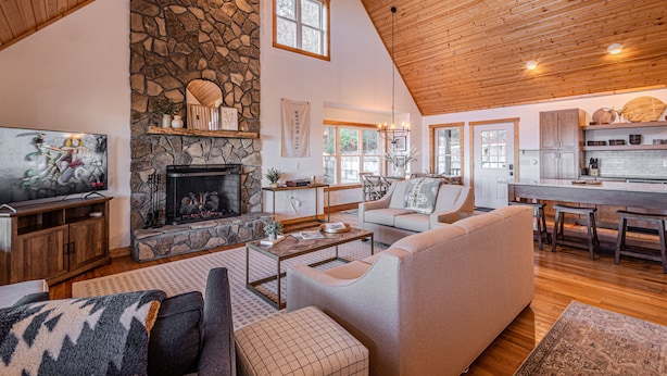 A spacious living area with a stone fireplace, wooden ceiling, and a kitchen area with bar stools.