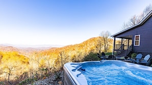 A hot tub with a view of a mountainous landscape, a deck with chairs, and a house with a porch.