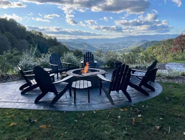 A circular outdoor seating area with a fire pit, surrounded by Adirondack chairs, set against a backdrop of a mountainous landscape.