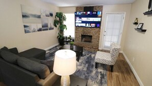 Een woonkamer met een open haard, een aan de muur bevestigde televisie, een bank, een fauteuil en een lamp.