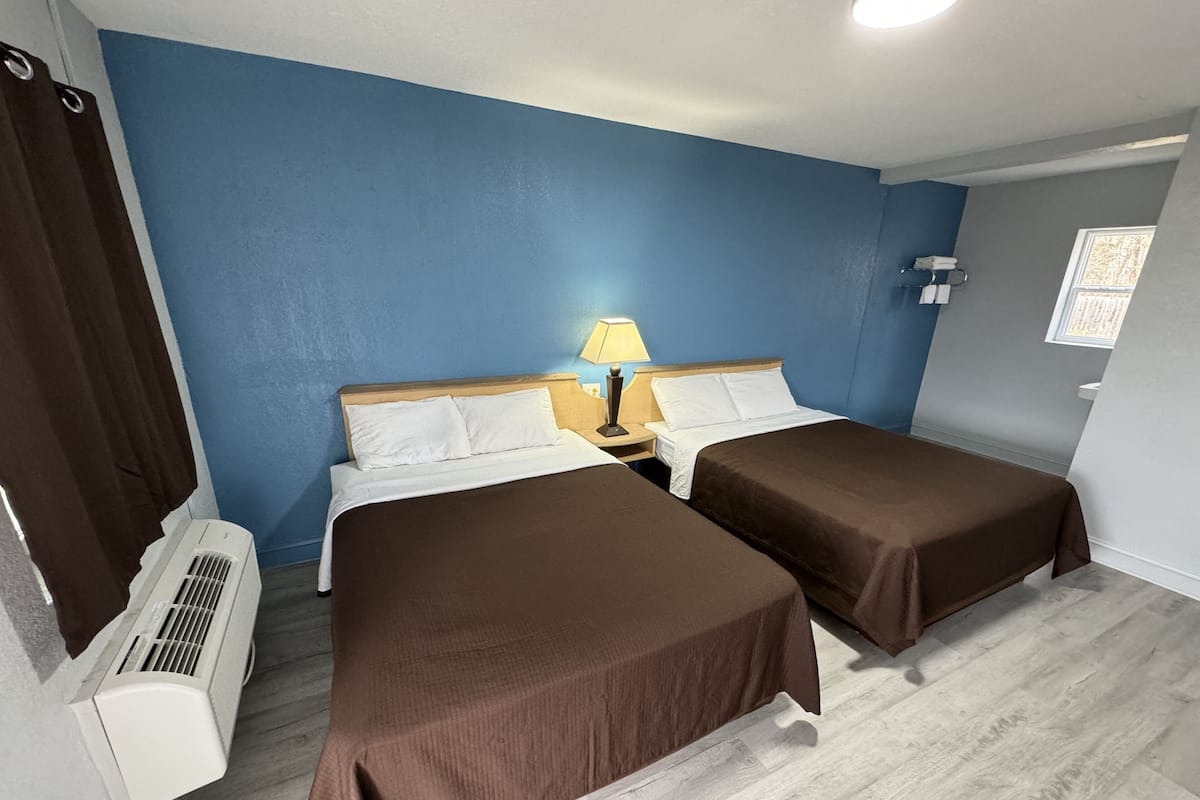 A room with two beds, a nightstand with a lamp, a window, and an air conditioning unit.