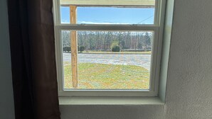 A room with a window overlooking a grassy area and trees, and a wall-mounted air conditioning unit below the window. - Budget Inn (Thomasville)