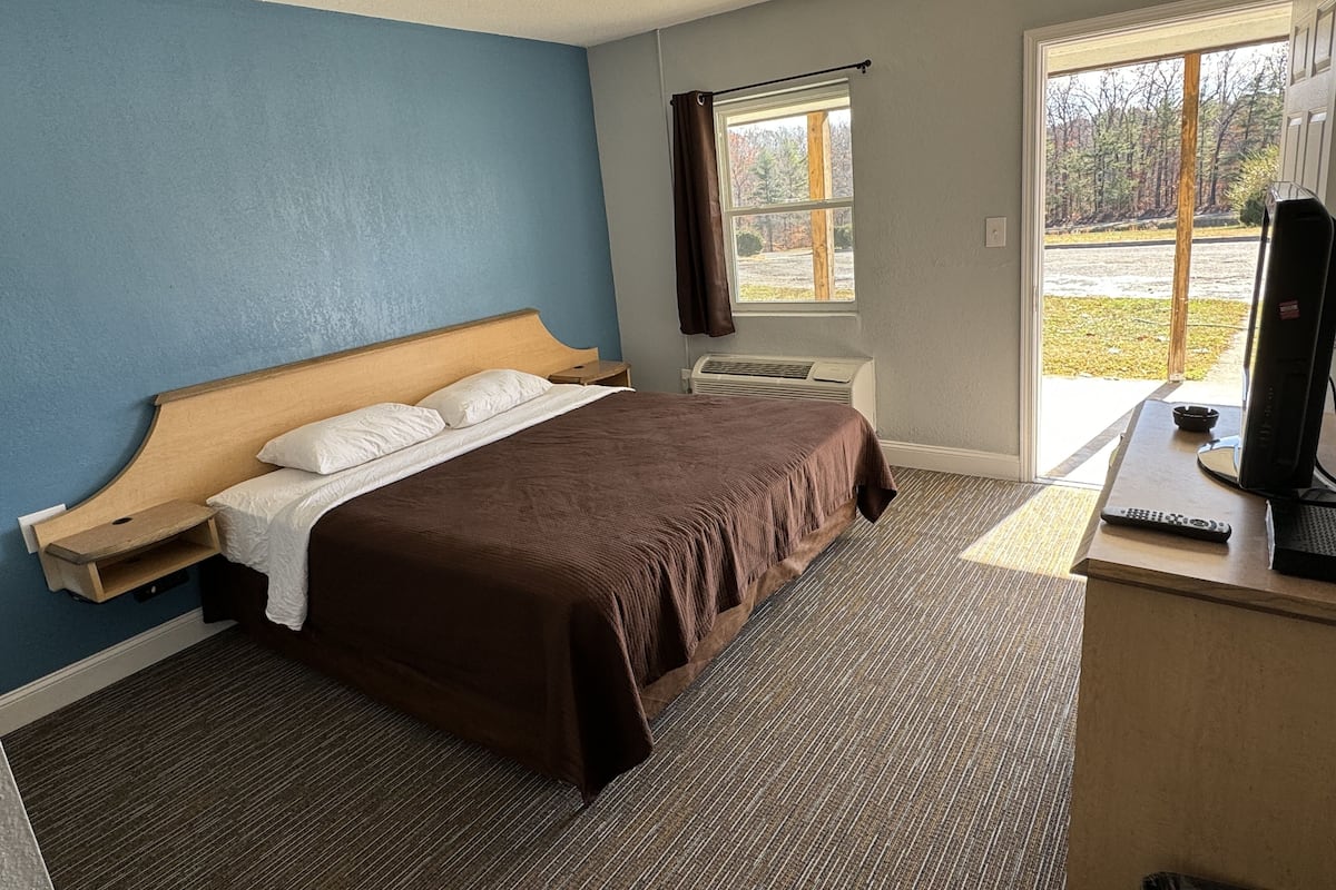 A hotel room with a large bed, a desk with a computer, a window with a view of trees, and a door leading outside.