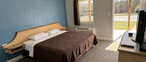 A hotel room with a large bed, a desk with a computer, a window with a view of trees, and a door leading outside.