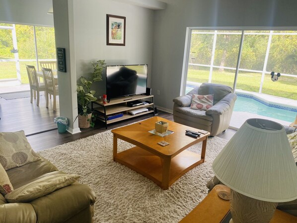 Una sala de estar con televisión, una mesa de centro, un sofá, una silla y una lámpara. Se ve una ventana con vista a una piscina y follaje al exterior.