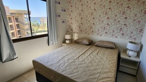 A bedroom with a bed, a nightstand, a lamp, and a view of a building outside. - Aldeia do Lago 306H (Caldas Novas)