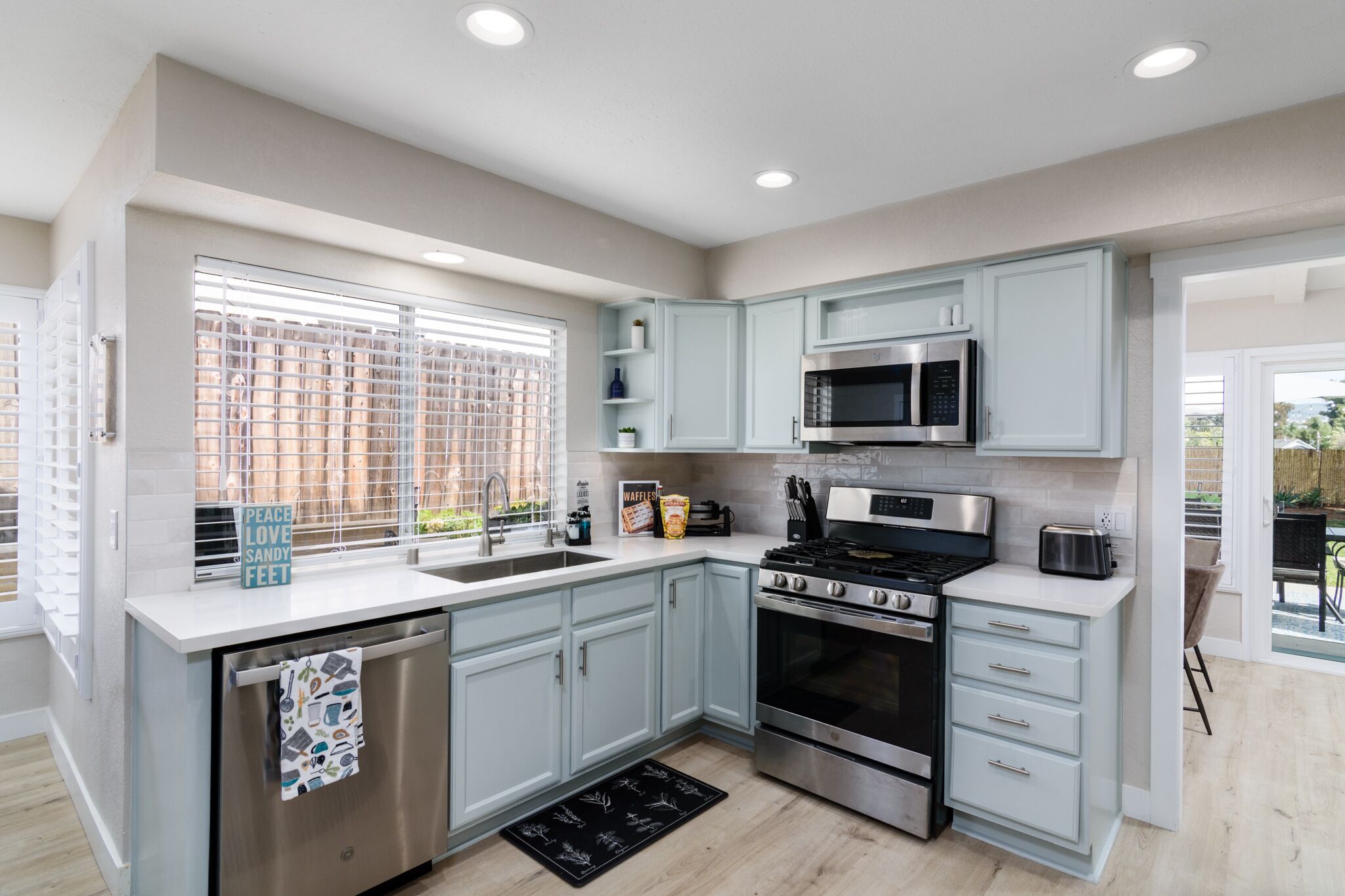 A modern kitchen with light blue cabinets, stainless steel appliances, and a large window with blinds.