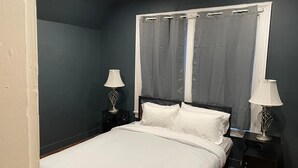 Quarto com cama, duas mesas de cabeceira com luminárias, uma janela com cortinas e uma parede escura.
