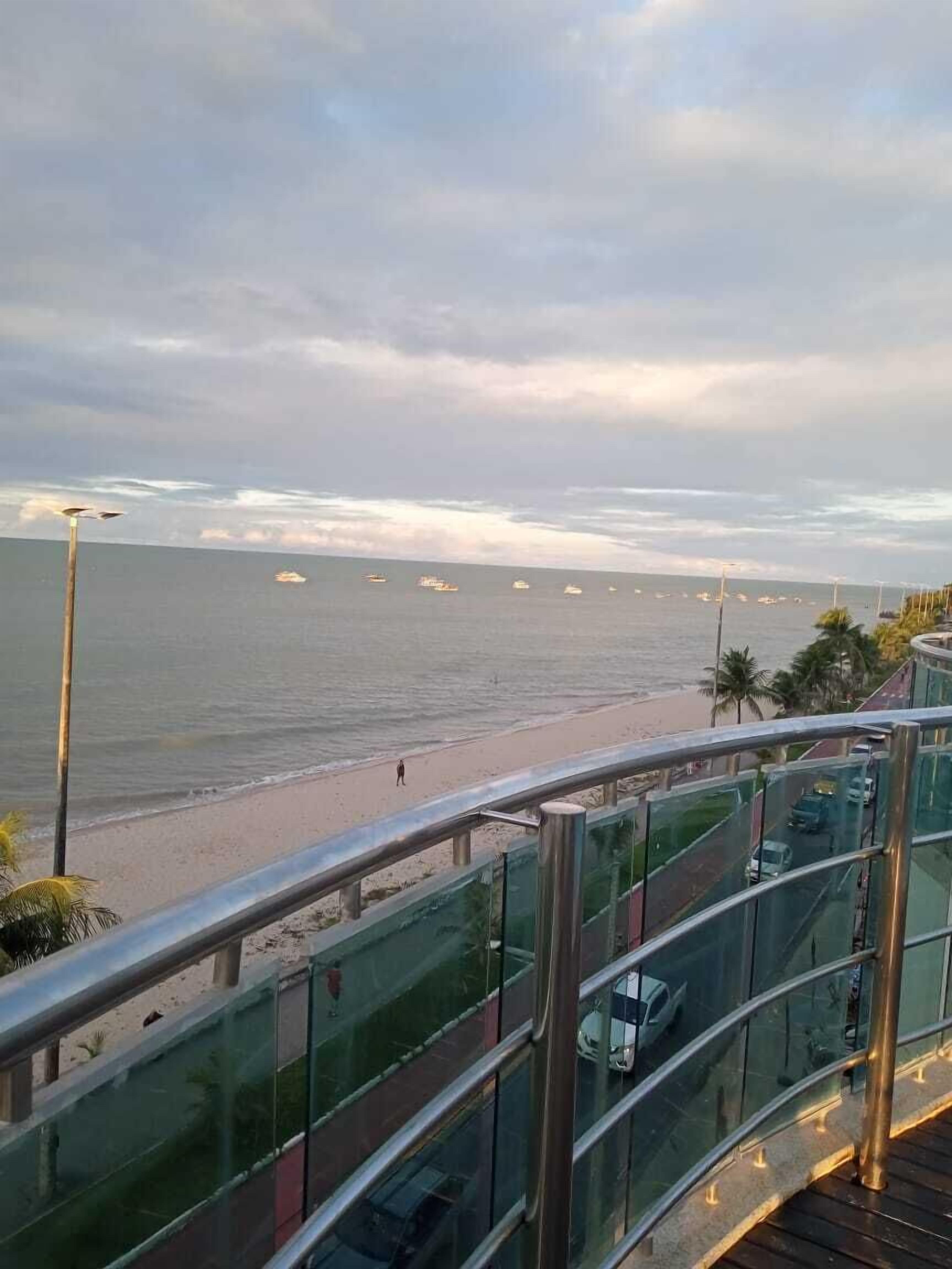 A coastal view from a balcony with a glass railing, overlooking a beach and the sea.
