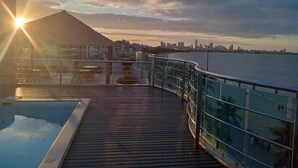 A wooden deck with a glass railing overlooking a body of water and a city skyline. - Manaira Flat (João Pessoa)