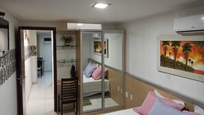 A bedroom with a bed, a chair, a desk, a mirror, and a painting on the wall. - Manaira Flat (João Pessoa)